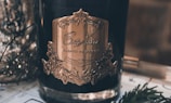 A close-up of a luxury candle labeled as 'Cote Noire Parfum de Maison' with an ornate, gold-embossed label. The candle is set amidst a cozy, dimly lit setting with decorative elements around it, including some evergreen branches and glittering ornaments in soft focus in the background.