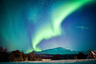A vibrant aurora borealis illuminates the night sky in shades of green and blue over a snowy landscape. The mountains in the background are partially covered in snow, and a traditional tent structure can be seen to the right. Trees in the foreground add depth to the serene scene.