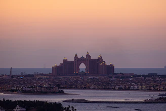 An expansive view of a coastal cityscape dominated by a large, iconic hotel with distinctive architecture featuring multiple towers and arches. The orange-pink hues of the sky suggest it is either sunrise or sunset. The foreground is a body of water with small islands and palm trees, while the background reveals a calm sea merging with the sky.