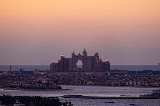 An expansive view of a coastal cityscape dominated by a large, iconic hotel with distinctive architecture featuring multiple towers and arches. The orange-pink hues of the sky suggest it is either sunrise or sunset. The foreground is a body of water with small islands and palm trees, while the background reveals a calm sea merging with the sky.