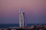 A sail-shaped luxury hotel stands majestically by the sea under a serene, gradient-colored sky. The structure is surrounded by water, with low-rise buildings in the foreground and a vast ocean fading into the horizon.