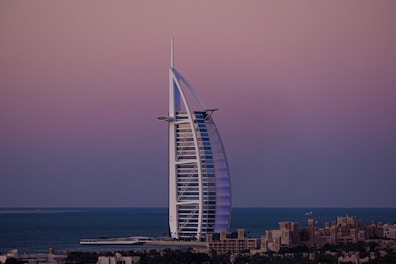A sail-shaped luxury hotel stands majestically by the sea under a serene, gradient-colored sky. The structure is surrounded by water, with low-rise buildings in the foreground and a vast ocean fading into the horizon.