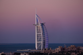 A sail-shaped luxury hotel stands majestically by the sea under a serene, gradient-colored sky. The structure is surrounded by water, with low-rise buildings in the foreground and a vast ocean fading into the horizon.