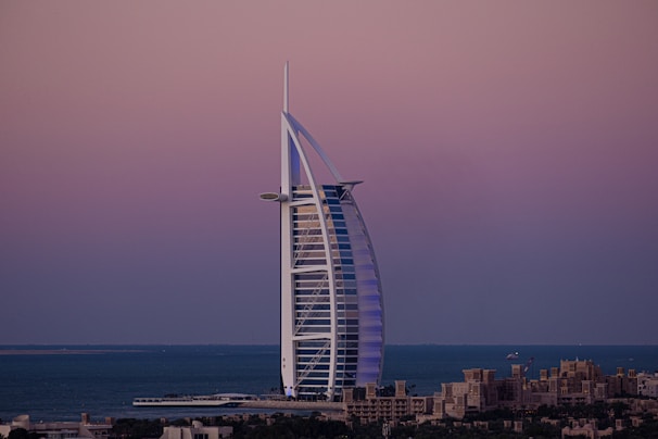 A sail-shaped luxury hotel stands majestically by the sea under a serene, gradient-colored sky. The structure is surrounded by water, with low-rise buildings in the foreground and a vast ocean fading into the horizon.