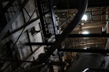An industrial setting with metal grates, a large pipe, and various structural components. The scene is dimly lit, casting shadows and creating a moody, mechanical atmosphere.