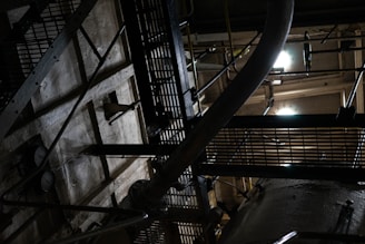 An industrial setting with metal grates, a large pipe, and various structural components. The scene is dimly lit, casting shadows and creating a moody, mechanical atmosphere.