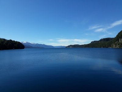 A tranquil lake reflecting the surrounding forest and distant mountains under a clear blue sky.