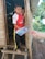 Two children stand at the entrance of a rustic wooden hut. The child at the front, wearing a red sports jersey, is receiving a book from an outstretched hand. The second child peeks from behind, partially hidden. The surrounding area includes a wooden structure, greenery, and a chicken wandering nearby.