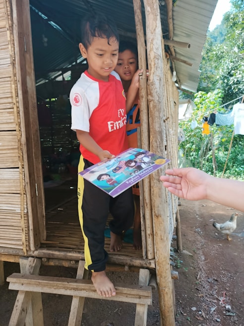 Two children stand at the entrance of a rustic wooden hut. The child at the front, wearing a red sports jersey, is receiving a book from an outstretched hand. The second child peeks from behind, partially hidden. The surrounding area includes a wooden structure, greenery, and a chicken wandering nearby.