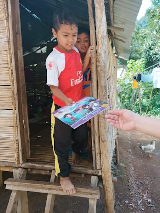 Two children stand at the entrance of a rustic wooden hut. The child at the front, wearing a red sports jersey, is receiving a book from an outstretched hand. The second child peeks from behind, partially hidden. The surrounding area includes a wooden structure, greenery, and a chicken wandering nearby.