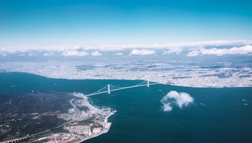 Aerial view of a large bridge spanning across a body of water, connecting two landmasses with a city visible in the background. The landscape is partly covered by clouds, with the water appearing dark blue and the sky light blue, dotted with white clouds.