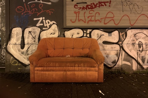 A worn-out orange-brown couch is placed against a graffiti-covered wall in an urban setting. The wall features numerous graffiti tags and stylized writings in various colors. The ground is a dark, paved surface with some dirt and small debris scattered around.