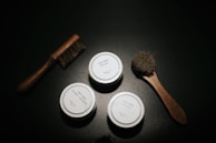 A dark surface with wooden-handled brushes and small round containers labeled 'Polish' and 'Clean + Hydrate' placed aesthetically. The scene has a minimalistic and organized arrangement.