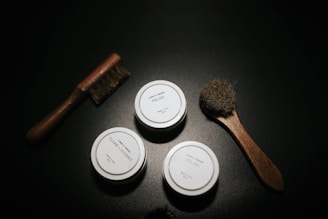A dark surface with wooden-handled brushes and small round containers labeled 'Polish' and 'Clean + Hydrate' placed aesthetically. The scene has a minimalistic and organized arrangement.