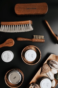 A collection of grooming products is displayed, including various brushes with wooden handles and soft bristles. There are also containers labeled as 'CLEAN + HYDRATE,' 'POLISH,' and 'WAX,' along with wooden bowls holding these products. Natural elements such as nuts or seeds are arranged on a wooden tray.