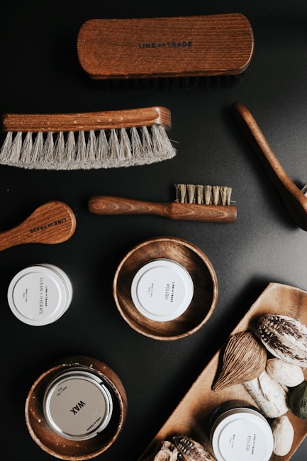 A collection of grooming products is displayed, including various brushes with wooden handles and soft bristles. There are also containers labeled as 'CLEAN + HYDRATE,' 'POLISH,' and 'WAX,' along with wooden bowls holding these products. Natural elements such as nuts or seeds are arranged on a wooden tray.