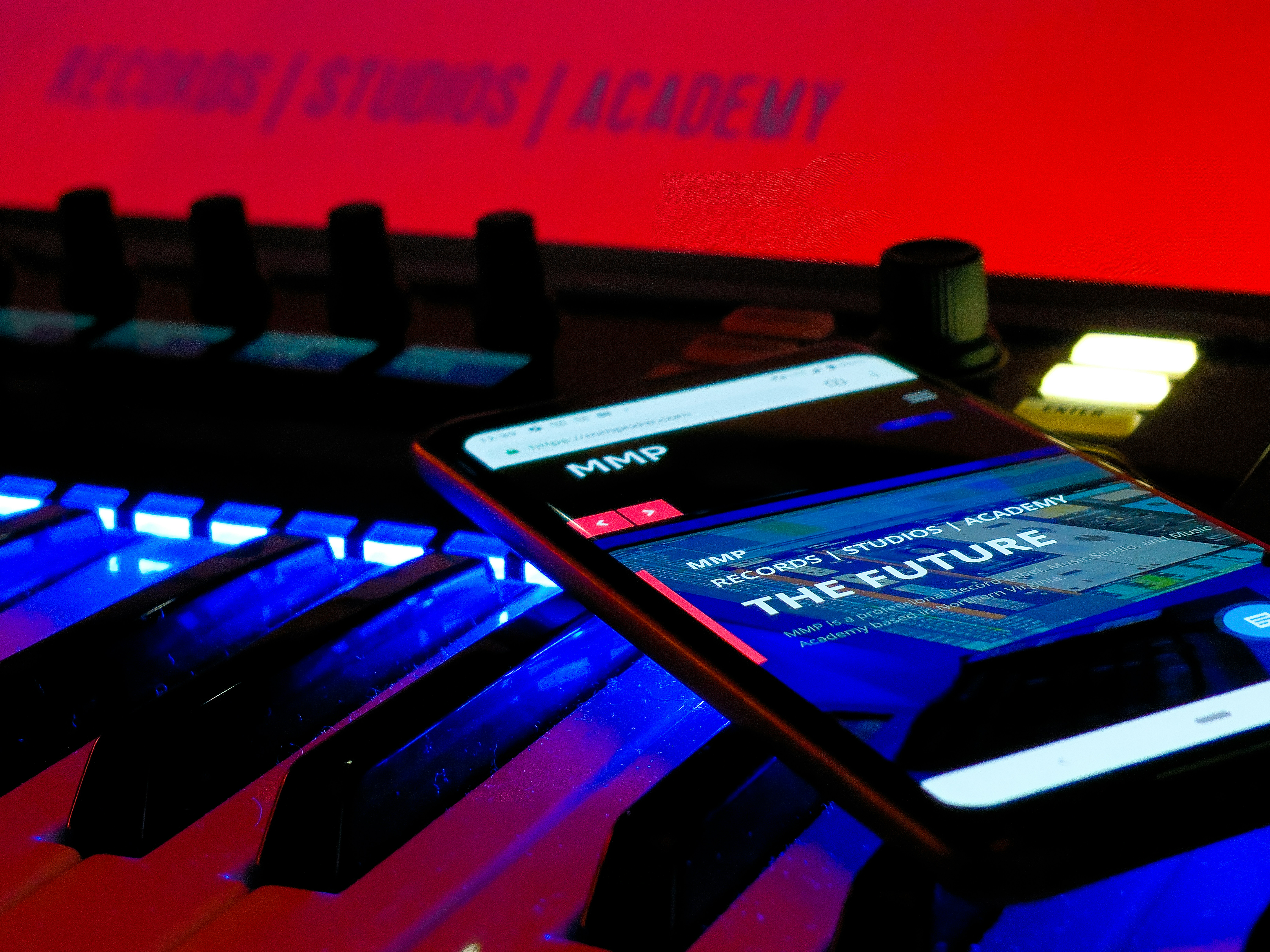 A smartphone displays a web page with the text 'THE FUTURE' and 'RECORDS / STUDIOS / ACADEMY' while placed on an illuminated music keyboard. The background features a bright red sign that also reads 'RECORDS / STUDIOS / ACADEMY'. The environment has a futuristic and technologically advanced feel.
