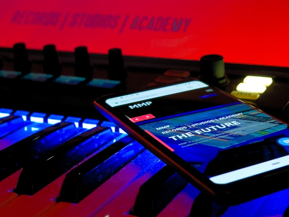 A smartphone displays a web page with the text 'THE FUTURE' and 'RECORDS / STUDIOS / ACADEMY' while placed on an illuminated music keyboard. The background features a bright red sign that also reads 'RECORDS / STUDIOS / ACADEMY'. The environment has a futuristic and technologically advanced feel.