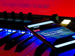A smartphone displays a web page with the text 'THE FUTURE' and 'RECORDS / STUDIOS / ACADEMY' while placed on an illuminated music keyboard. The background features a bright red sign that also reads 'RECORDS / STUDIOS / ACADEMY'. The environment has a futuristic and technologically advanced feel.