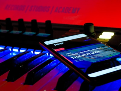 A smartphone displays a web page with the text 'THE FUTURE' and 'RECORDS / STUDIOS / ACADEMY' while placed on an illuminated music keyboard. The background features a bright red sign that also reads 'RECORDS / STUDIOS / ACADEMY'. The environment has a futuristic and technologically advanced feel.