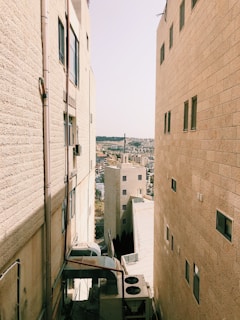 Tall, beige stone buildings form a narrow alleyway, with a view of a distant landscape dominated by a cross-topped building. The urban scenery is characterized by flat roofs and windows, with some air conditioning units and piping visible in the alley.