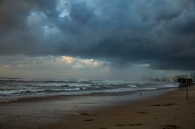 Dark, stormy clouds hang over a deserted beach with waves crashing against the shore. The skyline includes distant buildings partly obscured by mist, enhancing the dramatic and ominous atmosphere.
