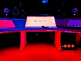 A modern music studio with vibrant red and blue lighting, featuring a large mixing console with a digital display showing the logo 'MMP'. Studio monitors and electronic equipment are visible around the console.