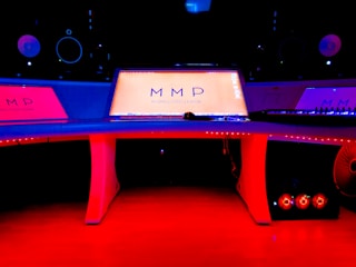 A modern music studio with vibrant red and blue lighting, featuring a large mixing console with a digital display showing the logo 'MMP'. Studio monitors and electronic equipment are visible around the console.