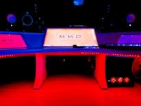 A modern music studio with vibrant red and blue lighting, featuring a large mixing console with a digital display showing the logo 'MMP'. Studio monitors and electronic equipment are visible around the console.