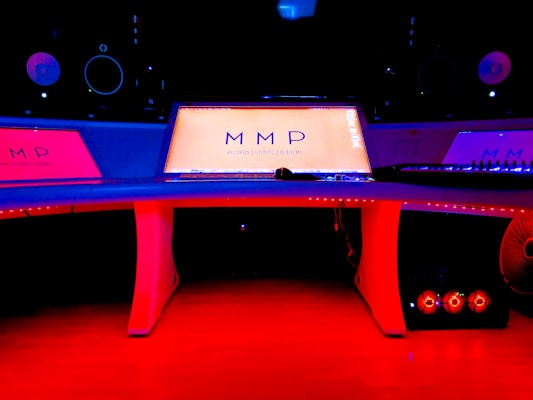 A modern music studio with vibrant red and blue lighting, featuring a large mixing console with a digital display showing the logo 'MMP'. Studio monitors and electronic equipment are visible around the console.