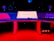 A modern music studio with vibrant red and blue lighting, featuring a large mixing console with a digital display showing the logo 'MMP'. Studio monitors and electronic equipment are visible around the console.