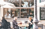 A cozy outdoor café setting features people seated at tables under large umbrellas, enjoying their meals or drinks. Inside, through a large window, individuals are engaged in reading and working on laptops. The window is surrounded by a blackboard with handwritten menu items and decorative books are visible on shelves.