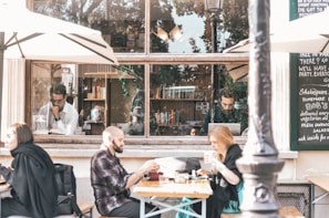 A cozy outdoor café setting features people seated at tables under large umbrellas, enjoying their meals or drinks. Inside, through a large window, individuals are engaged in reading and working on laptops. The window is surrounded by a blackboard with handwritten menu items and decorative books are visible on shelves.