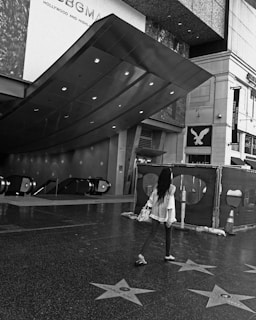 A black and white scene of a person walking along the Hollywood Walk of Fame, with stars embedded in the pavement. An overhead modern canopy and escalators are visible, along with a fenced-off area nearby. Nearby buildings bear signage for businesses.