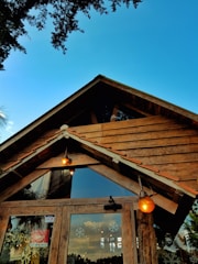 A rustic wooden building with a triangular roof, featuring two warm-toned outdoor lights and a glass entrance door. Snowflake decals can be seen on the glass, and the sky is clear blue with minimal trees in the background.