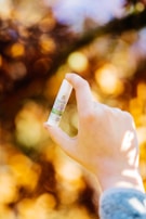 A hand holding a small tube of hemp oil balm with a blurred background of autumn leaves in shades of orange and yellow.