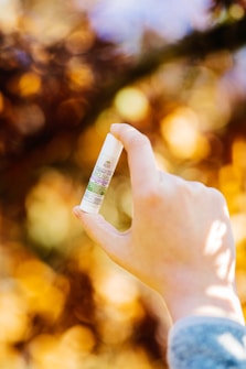 A hand holding a small tube of hemp oil balm with a blurred background of autumn leaves in shades of orange and yellow.