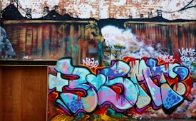 The image features a vibrant and colorful graffiti mural on a brick wall. The artwork incorporates a variety of bold colors and abstract shapes. The background includes darker shades with what appears to be a cityscape silhouette, while the foreground is dominated by vivid graffiti letters.