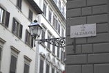A street scene featuring classic European architecture with a focus on beige and off-white buildings adorned with numerous windows and shutters. A vintage-style streetlamp is mounted on a decorative iron bracket extending from a wall. A street sign reads 'Via dei Calzaiuoli,' indicating an Italian location.