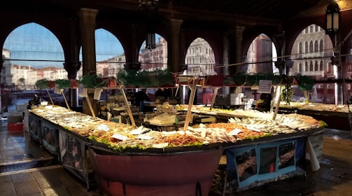 A bustling indoor seafood market with a large display of fresh fish and seafood on ice, surrounded by decorative arches and dim lighting. The architecture has an old-world charm with intricate stonework and a view of historic buildings through the windows.