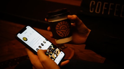 A person holds a smartphone displaying a social media profile page while simultaneously holding a cup of coffee in the other hand. The coffee cup has a brown sleeve with a design printed on it. The background is dimly lit, setting a warm and cozy atmosphere.