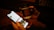A person holds a smartphone displaying a social media profile page while simultaneously holding a cup of coffee in the other hand. The coffee cup has a brown sleeve with a design printed on it. The background is dimly lit, setting a warm and cozy atmosphere.