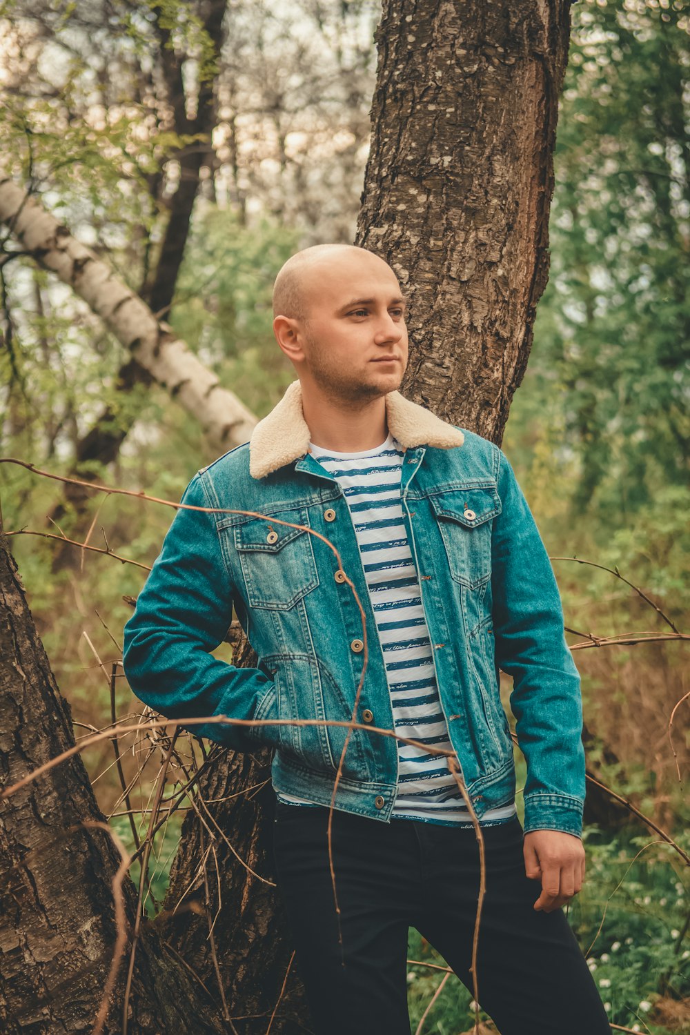 man wearing blue denim jacket and black pants standing beside tree during daytime