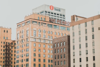 A cityscape featuring several multi-story brick and modern commercial buildings. The prominent building with a sign displaying 'THE PEABODY' is in the center, surrounded by other structures with varying architectural styles. High-rise buildings exhibit a mix of warm-toned bricks and neutral modern facades.