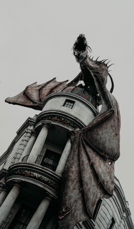 A large, menacing dragon perched atop a building with classical architectural features. The dragon's wings are spread wide, its mouth open in a roar, and sharp spikes line its body. The structure it rests on has tall columns and ornate details, giving a sense of grandeur.