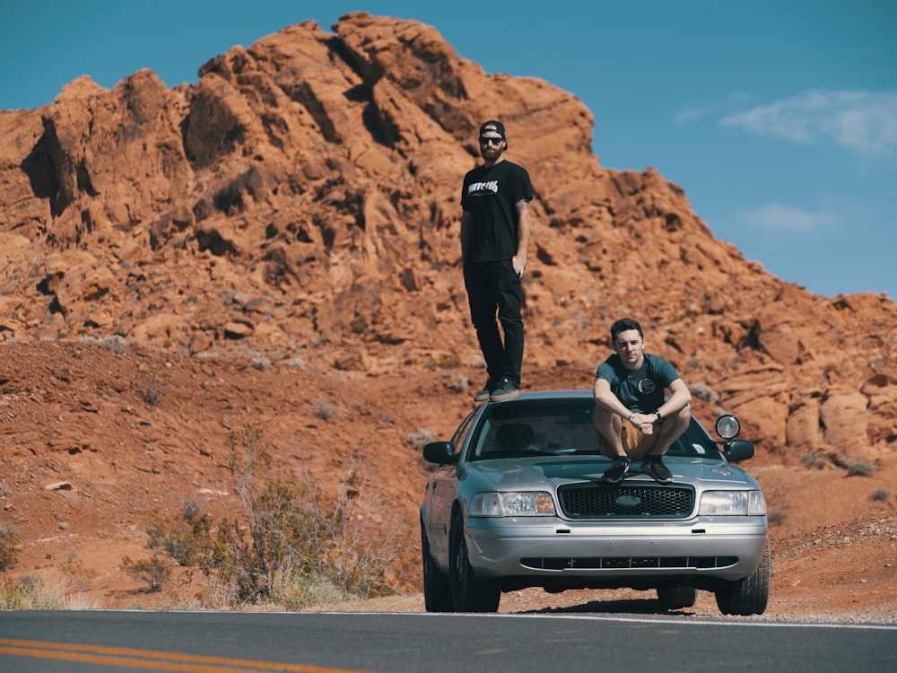 two men sitting and crouching on parked car beside road