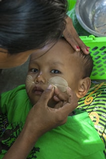 A child is lying down while an adult carefully applies a circular pattern with a light-colored substance on the child's cheeks. The child is wearing a bright green shirt, and there is a green basket and a metal bowl nearby. The patterned fabric beneath the child features colorful swirls.