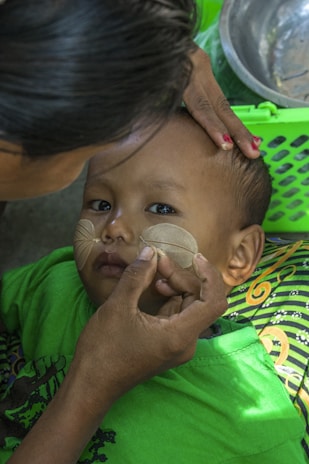 A child is lying down while an adult carefully applies a circular pattern with a light-colored substance on the child's cheeks. The child is wearing a bright green shirt, and there is a green basket and a metal bowl nearby. The patterned fabric beneath the child features colorful swirls.