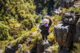 A person is zip-lining across a lush, green landscape, surrounded by dense foliage and rocky outcrops. The scene depicts adventure and nature, with the individual wearing a helmet and harness for safety.