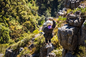 A person is zip-lining across a lush, green landscape, surrounded by dense foliage and rocky outcrops. The scene depicts adventure and nature, with the individual wearing a helmet and harness for safety.